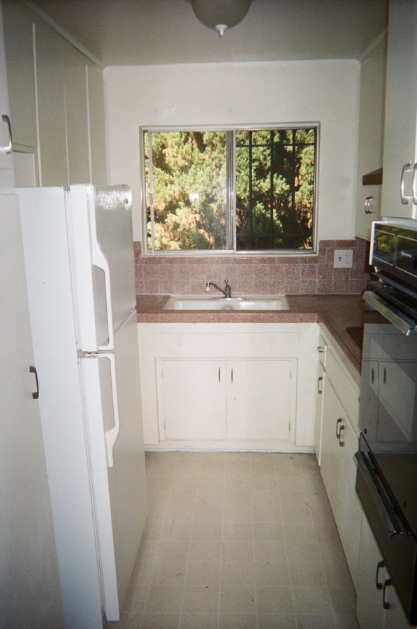 A small galley kitchen in our West Hollywood apartment, featuring white cabinetry and a pink-tiled countertop. A stainless steel sink is situated below a large window that overlooks lush green trees outside. A white refrigerator is on the left, with a built-in oven and stovetop visible on the right. The floor is a light cream-colored linoleum.