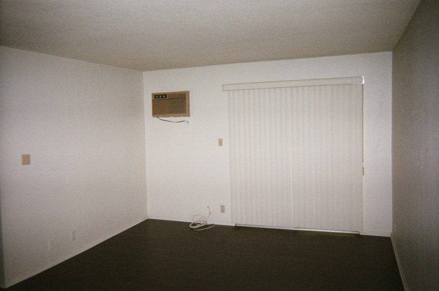 The now-empty living room in our West Hollywood apartment. A yellowed old wall-mounted air conditioning unit is to the left on the wall, while vertical blinds cover a sliding glass door on the right. The room has dark brown hardwood floors and minimal wall outlets visible near the floor. The lighting is even and natural, with no furniture remaining.