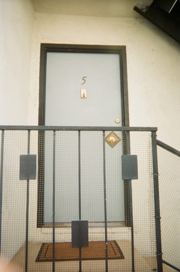 The door to our apartment, painted a pale blue with the number "5" displayed prominently. The door is framed by a black railing, and a simple brown doormat sits at the entrance. The stucco walls and subtle wear suggest the character of the building.
