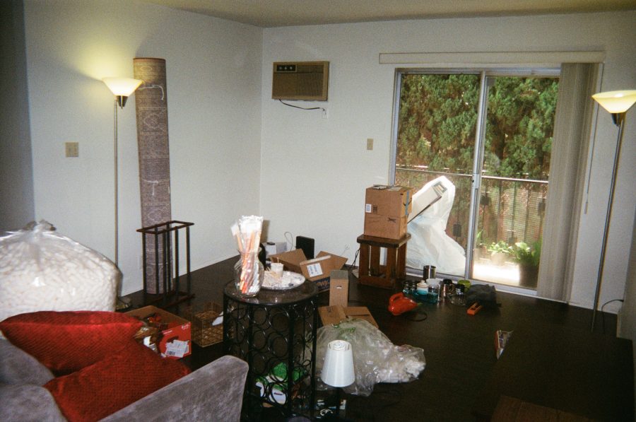 Another angle of the living room as we move out, showing the same gray couch, red cushion pillows, and packed boxes. The rug is tied up, and items like lamps, a side table, and loose household belongings are visible. Sliding doors open onto a patio lined with greenery.