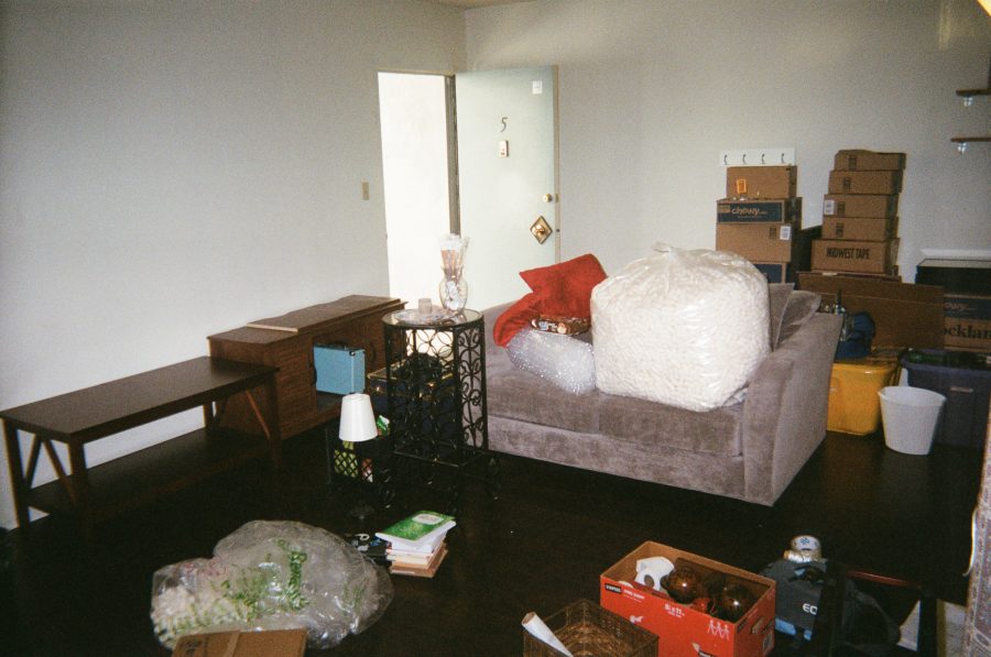 Interior of our West Hollywood apartment as we pack and prepare to move out. The living room features a gray couch with red throw pillows, bubble wrap, a massive bag of packing peanuts, and packed moving boxes. Furniture such as an end table with a decorative metal wine rack and a wooden shelf are scattered among the items.