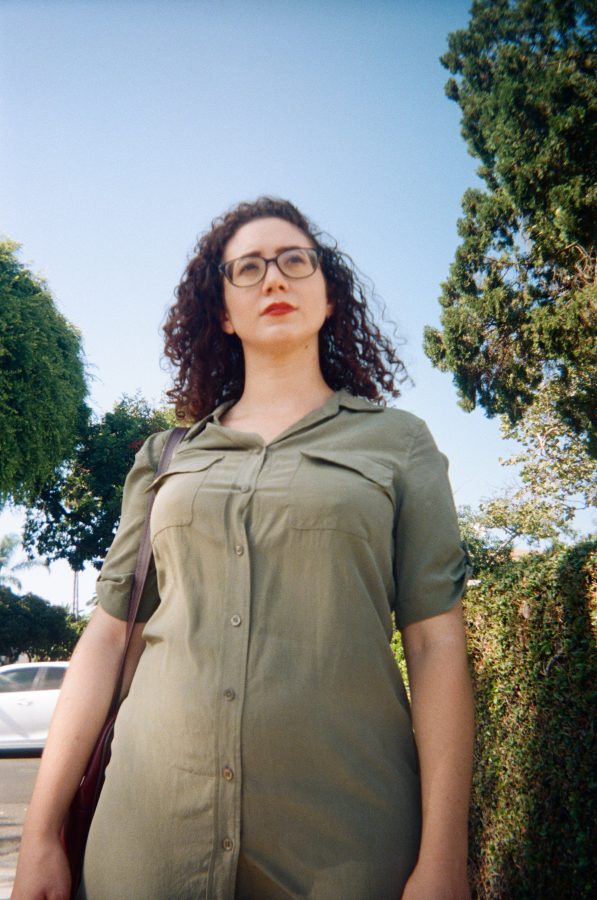 Marina standing boldly beneath a tree in West Hollywood, captured from a low angle. Her striking silhouette is accentuated by the flowing sage-green dress she wears. The backdrop includes large trees and a sunny Los Angeles neighborhood, with a soft focus on the surrounding architecture.
