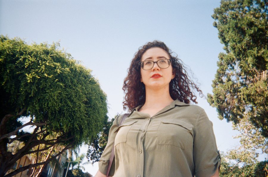 A close-up of Marina, framed by lush greenery and a bright blue Los Angeles sky. Her olive-green dress complements the natural backdrop, while her bold red lipstick and glasses draw attention to her expressive face. The trees surrounding her create a natural canopy, adding depth and dimension to the composition.