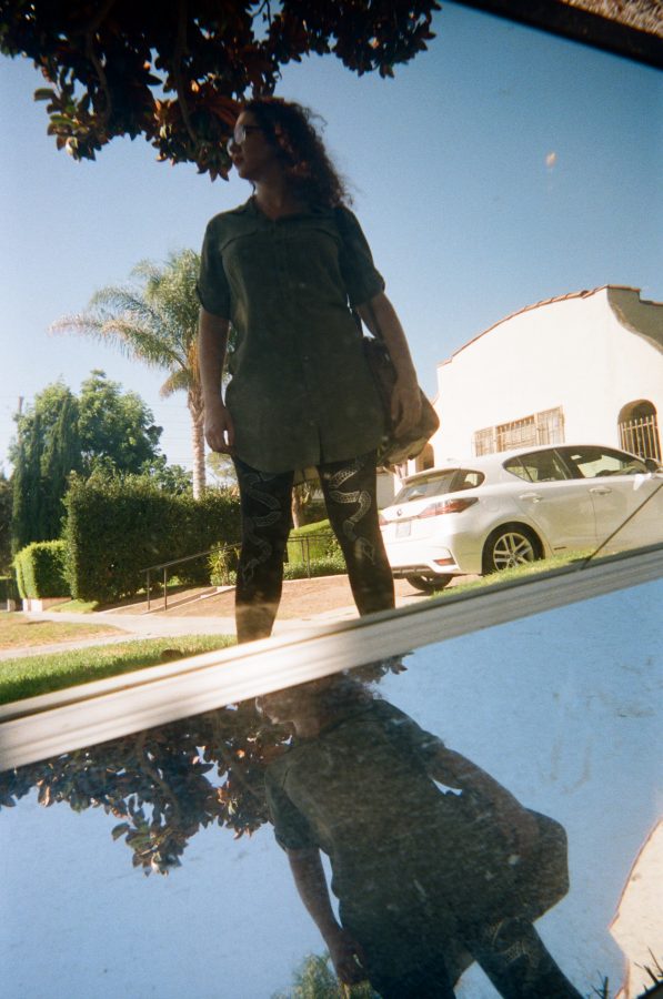 A creative shot of Marina reflected in a broken street mirror in West Hollywood. The reflection captures her standing confidently in her olive-green dress and snake-print leggings, framed by the tree-lined residential street and parked cars in the background. The cracked mirror adds a fragmented, artistic quality to the photo.