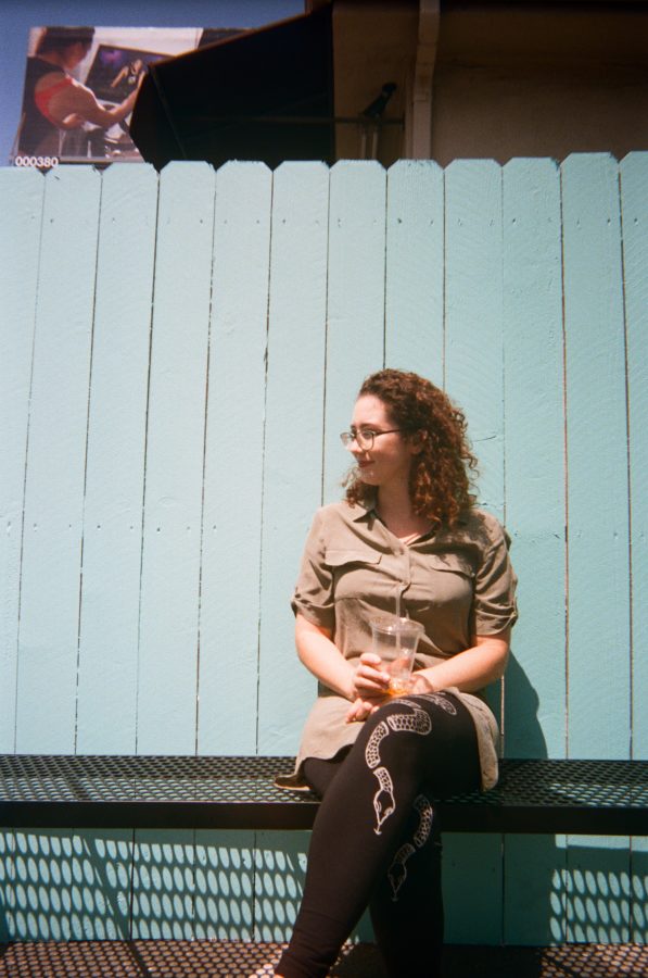Marina again in front of the turquoise fence, this time with a billboard partially visible in the background above her. She sits casually, looking off to the side with a thoughtful expression, her features highlighted by the sun. The contrasting bright and muted tones make this urban West Hollywood moment striking.