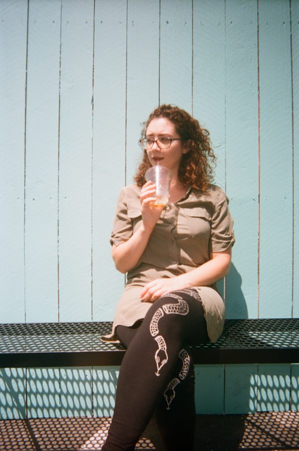 Marina sitting in front of a bright turquoise-painted wooden fence at Coffee Coffee. She holds an iced coffee in one hand, her snake-print leggings and olive-green dress standing out against the vibrant backdrop. Her curly hair catches the sunlight, and the soft shadows of the fence slats add texture to the scene.
