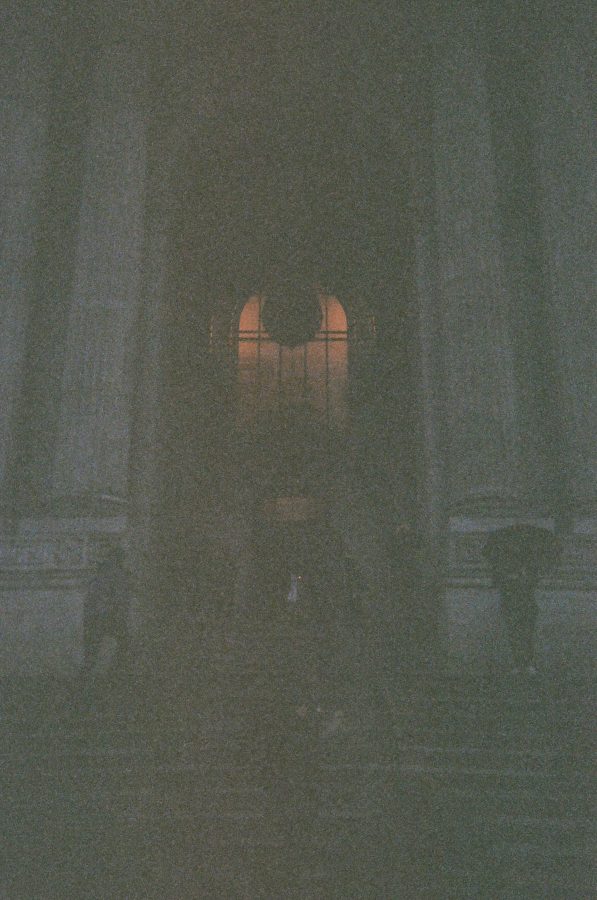 A closer view of the New York Public Library’s grand staircase and towering columns. The dim light emphasizes the imposing scale of the architecture, with the faint glow of a lantern visible through the central arched window. A few figures stand on the steps, bundled in coats and holding umbrellas, adding a sense of scale and life to the scene.
