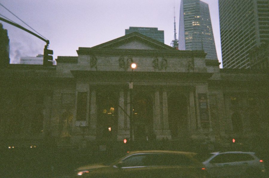 The exterior of the New York Public Library on Fifth Avenue is partially silhouetted against the early evening sky. The neoclassical facade features grand columns and intricate stone carvings, with soft light illuminating the arched windows. In the foreground, yellow taxis and other vehicles drive past, adding to the lively city atmosphere.