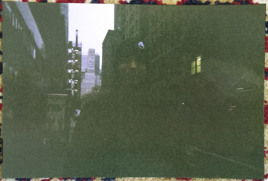 A physical photograph of the next glitched image, placed on a patterned rug. The photo shows me smiling in a black winter coat and a stocking cap, standing in a New York City street with skyscrapers in the back. The faint, blurred buildings in the background are barely discernible under the dim light.