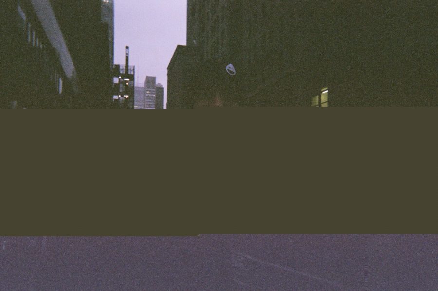 A glitched photo of me in New York City (the previous picture). The upper half shows me wearing a black winter coat and a stocking cap with a logo, with dimly lit buildings in the background. The middle of the photo is a solid brown strip where the image data is corrupted, and the bottom fades into a purple-tinted hue, creating a surreal, fragmented effect.