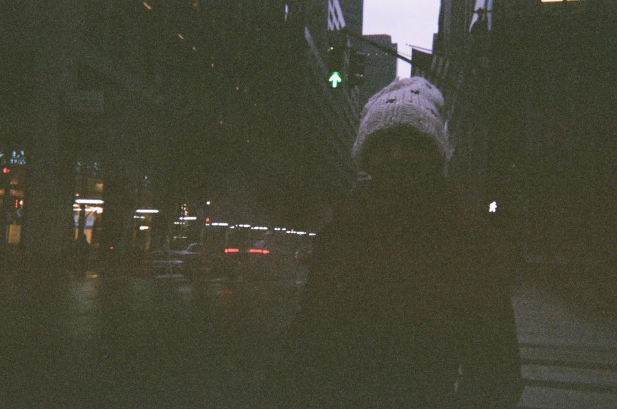 Alex stands on a New York City sidewalk, bundled up against the cold. She wears a thick knitted white beanie and a dark winter coat. The background is softly lit, with blurred city lights and the faint outline of buildings and traffic signals, emphasizing the urban setting.
