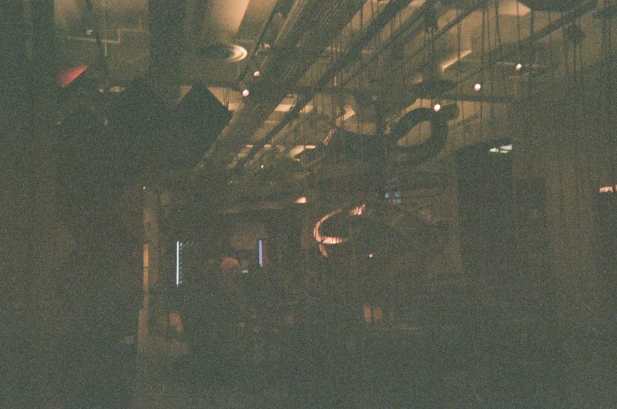 A dimly lit interior of the Museum of Mathematics features an eclectic mix of suspended, looping structures, cables, and spotlights. The ceiling is filled with exposed pipes and ducts, contributing to the industrial aesthetic. The ambient lighting creates a warm atmosphere, highlighting the museum's unconventional, interactive installations.