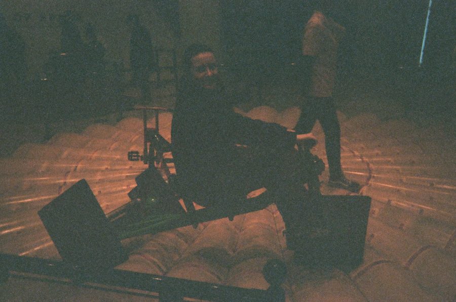 Alex sits on a quirky tricycle with square wheels, positioned on a floor designed with concentric tubular ridges at the Museum of Mathematics in New York City. The unique lighting casts warm, diffused shadows, emphasizing the geometry of the interactive exhibit. Behind Alex, a few other museum visitors and a large open space with exhibits are faintly visible.