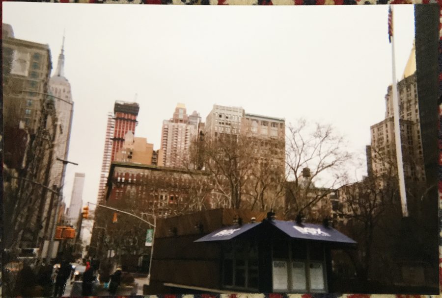 A physical photograph of New York City buildings, developed and resting on a colorful rug. The same skyline as the glitched image following this one is visible, with the Empire State Building and nearby structures standing out against a gray winter sky.