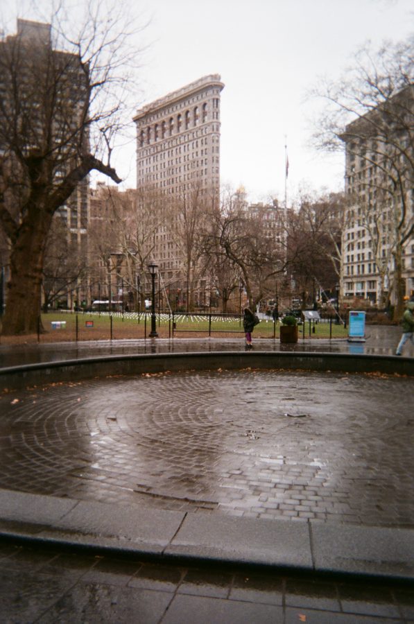 A view of the Flatiron District, showcasing the Flatiron Building's triangular architecture. A damp circular fountain sits in the foreground, its wet brick pattern glistening under soft light. Bare trees line the surrounding park, while pedestrians bundled in winter clothing walk nearby.
