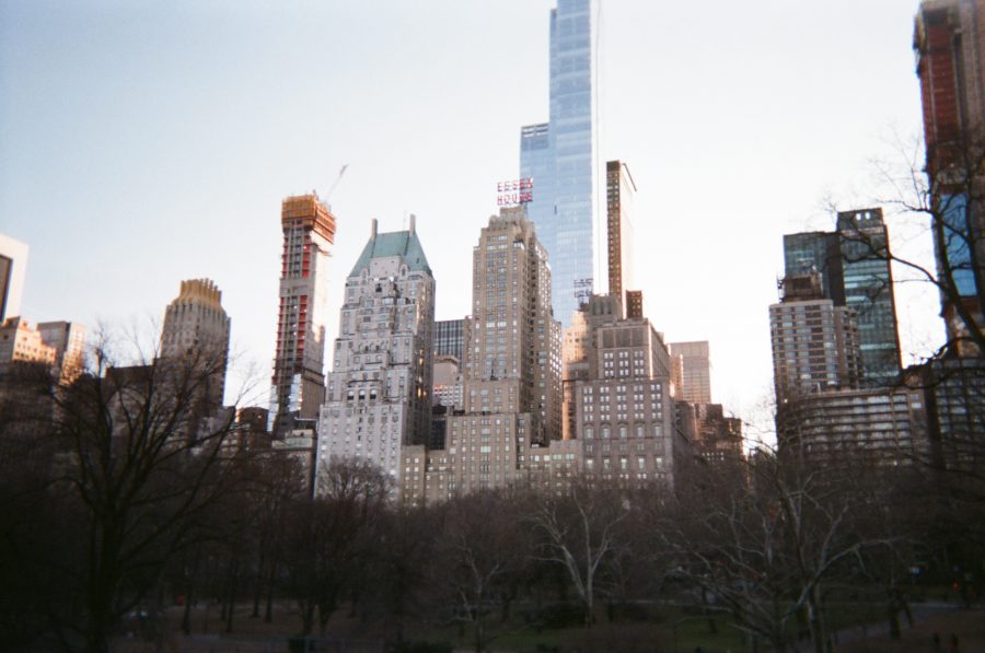 The Manhattan skyline as seen from Central Park. The mix of iconic high-rise buildings includes the Essex House sign atop a beige art-deco structure. Leafless winter trees dominate the foreground, contrasting the vertical geometry of the modern and historic architecture.
