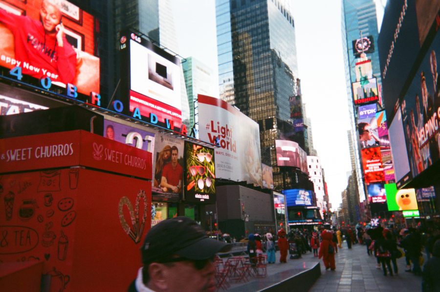 A street-level view of Times Square with an emphasis on the colorful chaos of signage. Billboards advertise products and messages, including a red Sweet Churros cart in the foreground and bright “#LightTheWorld” displays. The mix of modern architecture, streaming crowds, and street vendors brings the energy of New York City to life.