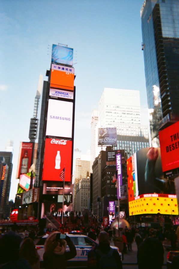 Times Square in New York City captured on a cold February day. Bright digital billboards tower over the scene, advertising brands like Samsung, Coca-Cola, and Xinhua News Agency. The iconic red steps are visible in the background, with people gathered around. The tall glass buildings reflect the light of the blue sky, while an NYPD vehicle is parked in the foreground, adding a sense of movement to the bustling area.