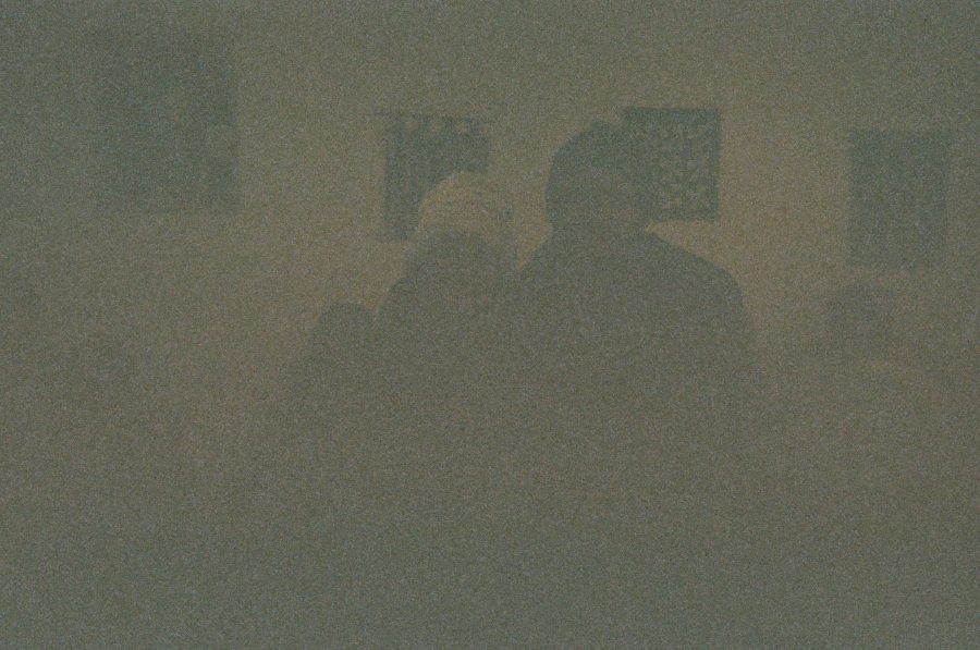 A heavily underexposed image of Alex and me, barely visible in a shadowy setting. Alex’s white stocking cap and dark jacket are faint outlines against a grainy, textured background. The environment is indistinct, with faint geometric shapes in the back that resemble picture frames or tapestries.