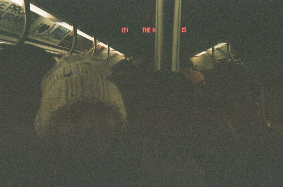 A tightly packed subway train on the F line in New York City. The dimly lit interior is crowded with passengers, with Alex visible wearing a white knitted stocking cap and smiling brightly. The overhead advertisements add small pops of color, and the LED display reads “F THE N.........IS" in red letters.