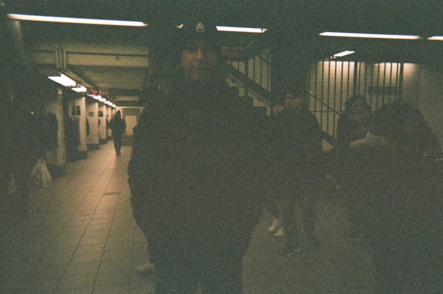 Me standing in a subway station wearing a red fluffy jacket and a black stocking cap with the Rumble Boxing logo. The background features tiled walls, staircases, and several people moving through the space. The fluorescent lighting adds a utilitarian feel, highlighting the subway’s bustling energy.