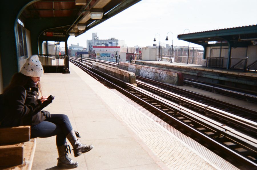Alex seated on a wooden bench at a Brooklyn MTA train station. She wears a black coat and a white knit hat, focused on her phone. The platform is open to the sky, with visible train tracks running parallel. A graffiti-covered wall and urban buildings form the distant backdrop.