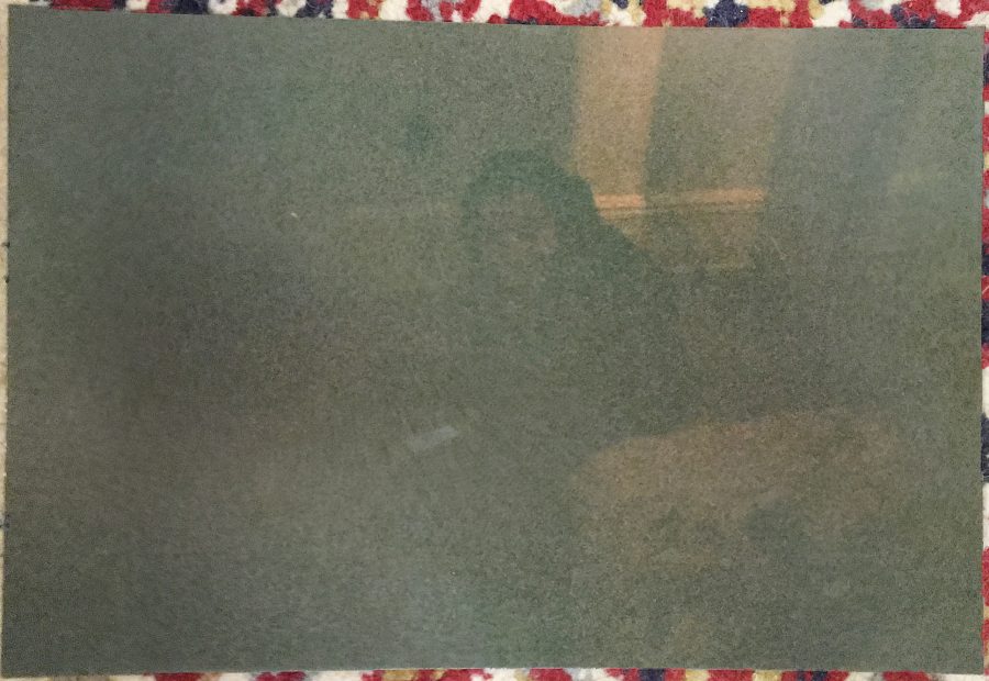 A developed photograph of Alex in a dimly lit location, placed on the same patterned rug as the mural photo. Alex is wearing dark clothing, and the lighting emphasizes the ambient shadows around them.