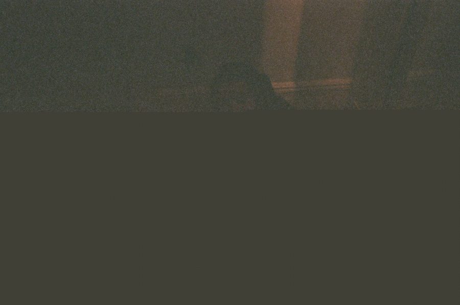A glitched digital image of Alex, with most of the bottom half obscured by a solid brown void. The upper portion hints at a dimly lit interior, with shadowy forms and warm tones. The distortion dominates the composition, giving it an abstract, otherworldly quality.