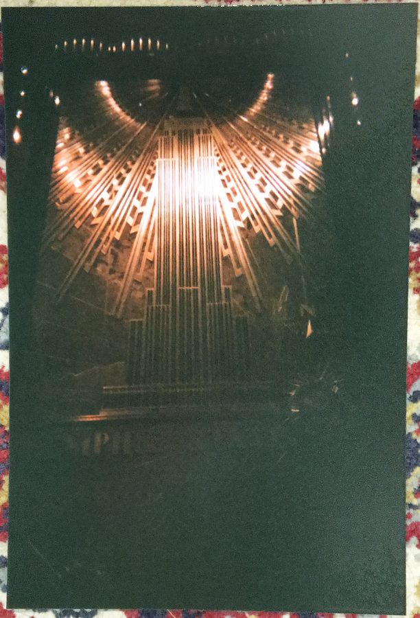 A photo of a developed physical photograph showing the interior art deco mural of the Empire State Building. The mural features glowing geometric patterns in bronze and gold hues, radiating upward. The physical photo rests on a colorful, textured rug with intricate red, blue, and beige patterns.