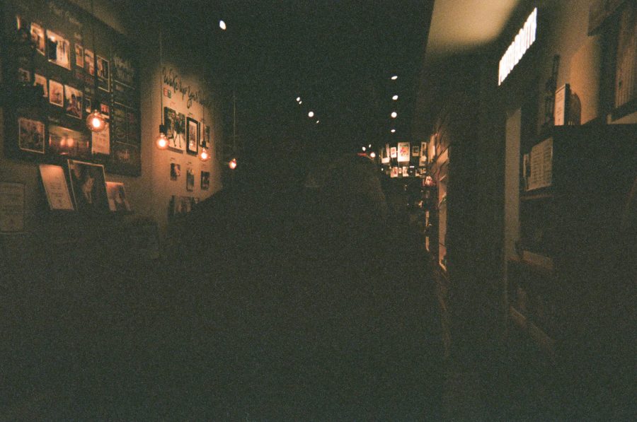 A dark, underexposed image of me and Alex inside the Wonder Photo Shop. The lighting highlights the faint outlines of picture frames on the wall, illuminated by small Edison-style bulbs. Shelving lines the right side, and the dimly lit walkway stretches into the background.
