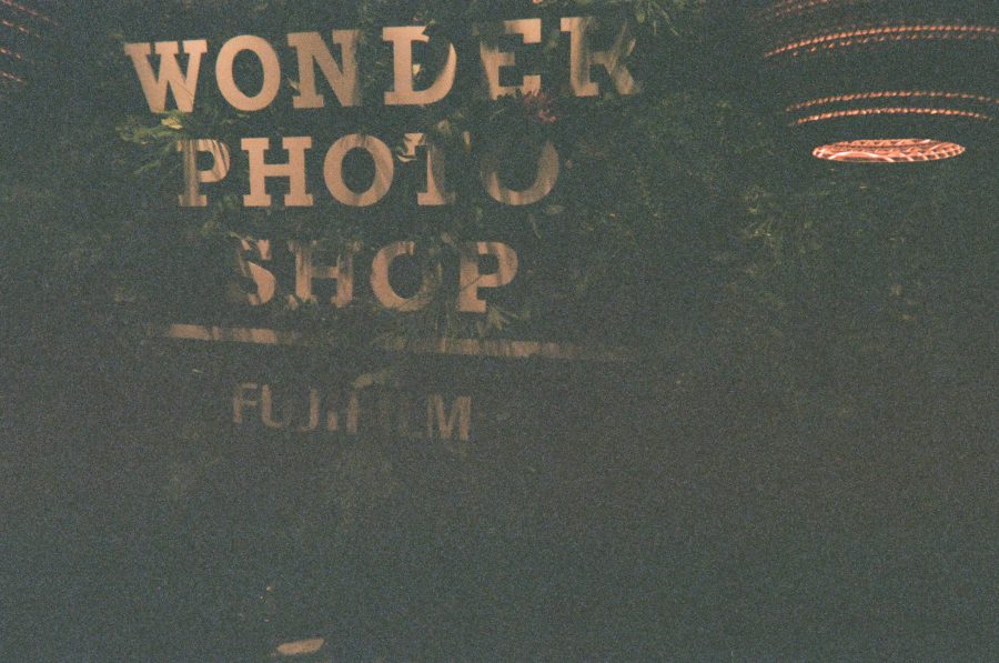 A storefront sign for "Wonder Photo Shop Fujifilm" partially obscured by lush, fake greenery. The white letters are large and bold, with a warm glow coming from circular hanging lamps on the sides. The scene is dimly lit, giving the sign a cozy, vintage feel.