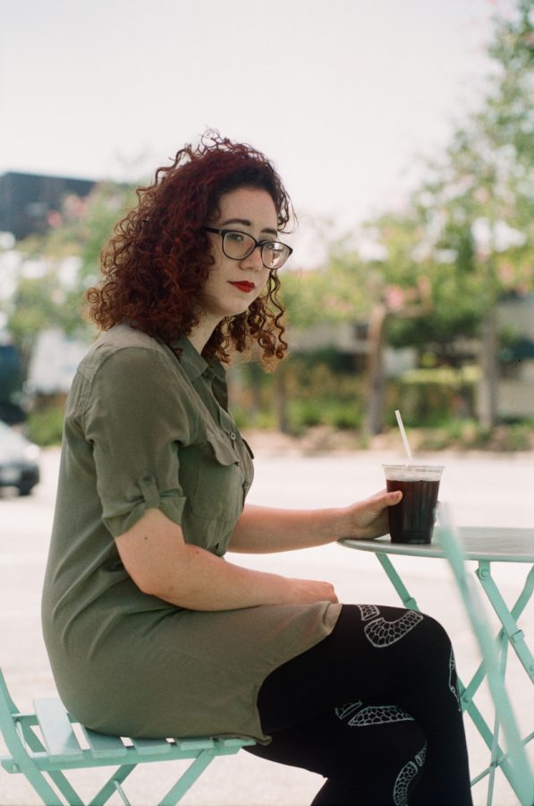 Marina sitting outdoors at a café called Coffee Coffee on Fairfax in West Hollywood. She wears a light olive-green shirt dress with her curly red hair framing her face, accented by dark glasses. Marina holds an iced coffee in her hand while sitting on a light turquoise metal chair at a matching table. The background includes soft greenery and blurred urban elements like a billboard and street trees, suggesting a sunny day in Los Angeles.