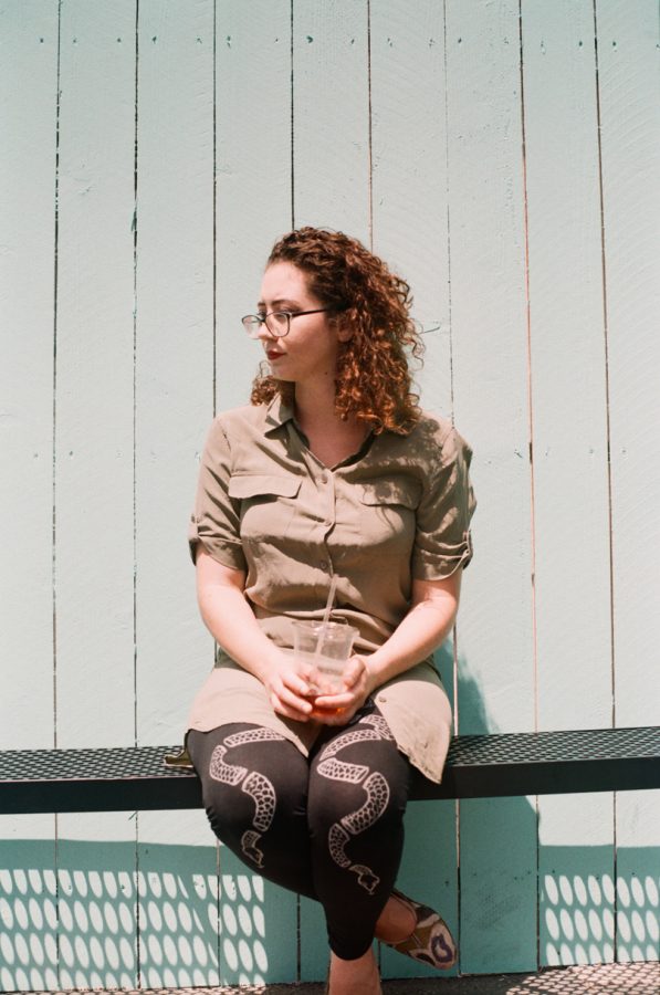 Marina on a sunny day, seated against a teal fence and holding an iced coffee cup. Her curly hair is tousled by the breeze, and the details of her outfit—a sage-green dress and snake-patterned leggings—are captured in natural light.