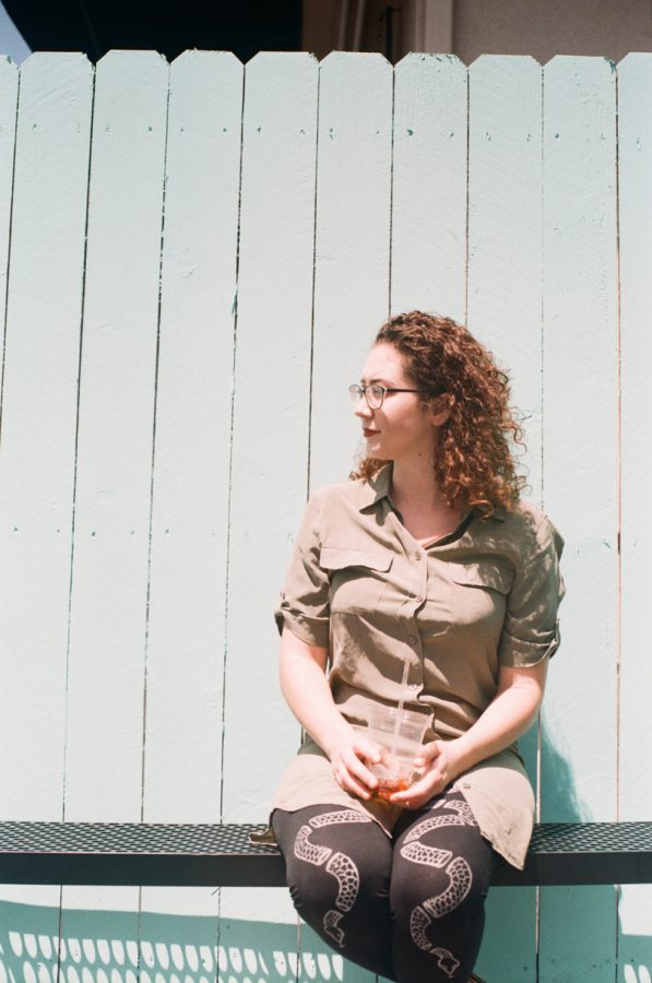 Marina seated outdoors, captured with a 35mm Petri film camera. She wears a sage-green dress and black leggings featuring snake designs, holding an iced coffee in a clear cup with a straw.