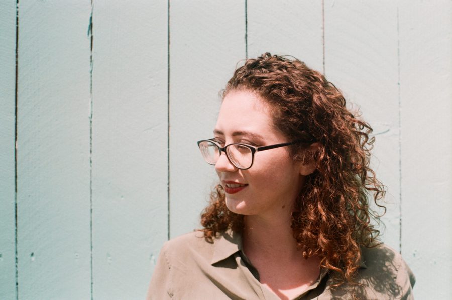 Close-up of Marina smiling softly, her curly auburn hair illuminated by sunlight. She wears glasses and a sage-green button-up dress, with a textured pale teal wooden fence as a backdrop.