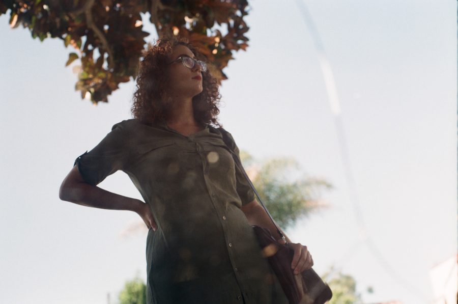 Marina reflected in a cracked street mirror in West Hollywood, standing confidently under a tree with lush green foliage. Her silhouette dominates the reflection, framed by palm trees, a parked white car, and a suburban Los Angeles street in the background.