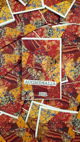 An artistic photo of the Augmenalia #03 zine issue laying face up on top of a pile of other Augmenalia #03 zine issues laying face down, showing off their marbled back covers, as though the issue is one in a sea of many.